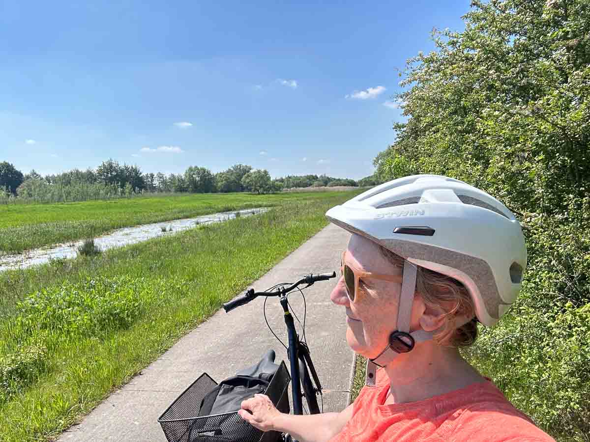Eine Person mit einem weißen Helm und einem orangefarbenen T-Shirt steht mit einem Fahrrad auf dem sonnigen Havel-Radweg an der Havel, neben grünen Feldern und Bäumen unter einem strahlend blauen Himmel.