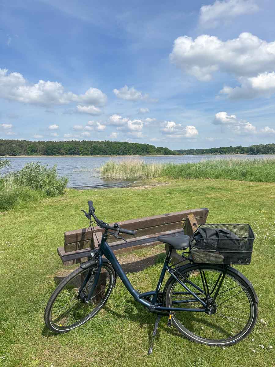 Neben einer Holzbank auf einer Rasenfläche in der Nähe des Hohennauener-Ferchesarer Sees im Westhavelland steht ein schwarzes Fahrrad; am Ufer wächst Schilf, und im Hintergrund erstreckt sich eine bewaldete Landschaft unter einem Himmel mit Schäfchenwolken.
