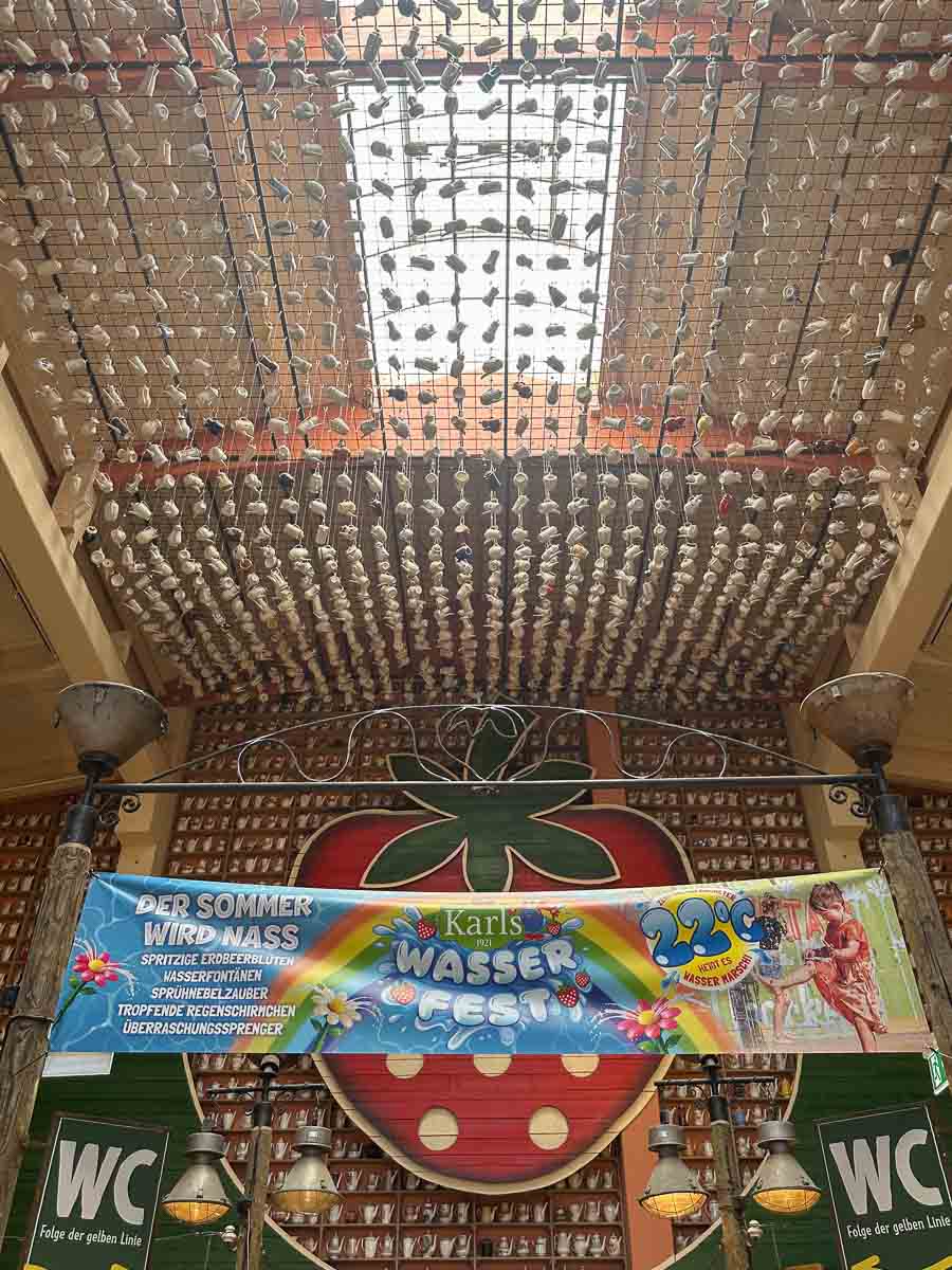 Ein dekorativ gestalteter Innenbereich in Karls Erleebnishof Elstal im Havelland besticht durch eine mit Kaffeekannen behangene Decke, ein großes Wandbild mit Erdbeeren und ein farbenfrohes Banner, das für ein Wasserfest wirbt. Unten sind auf beiden Seiten WC-Schilder zu sehen.