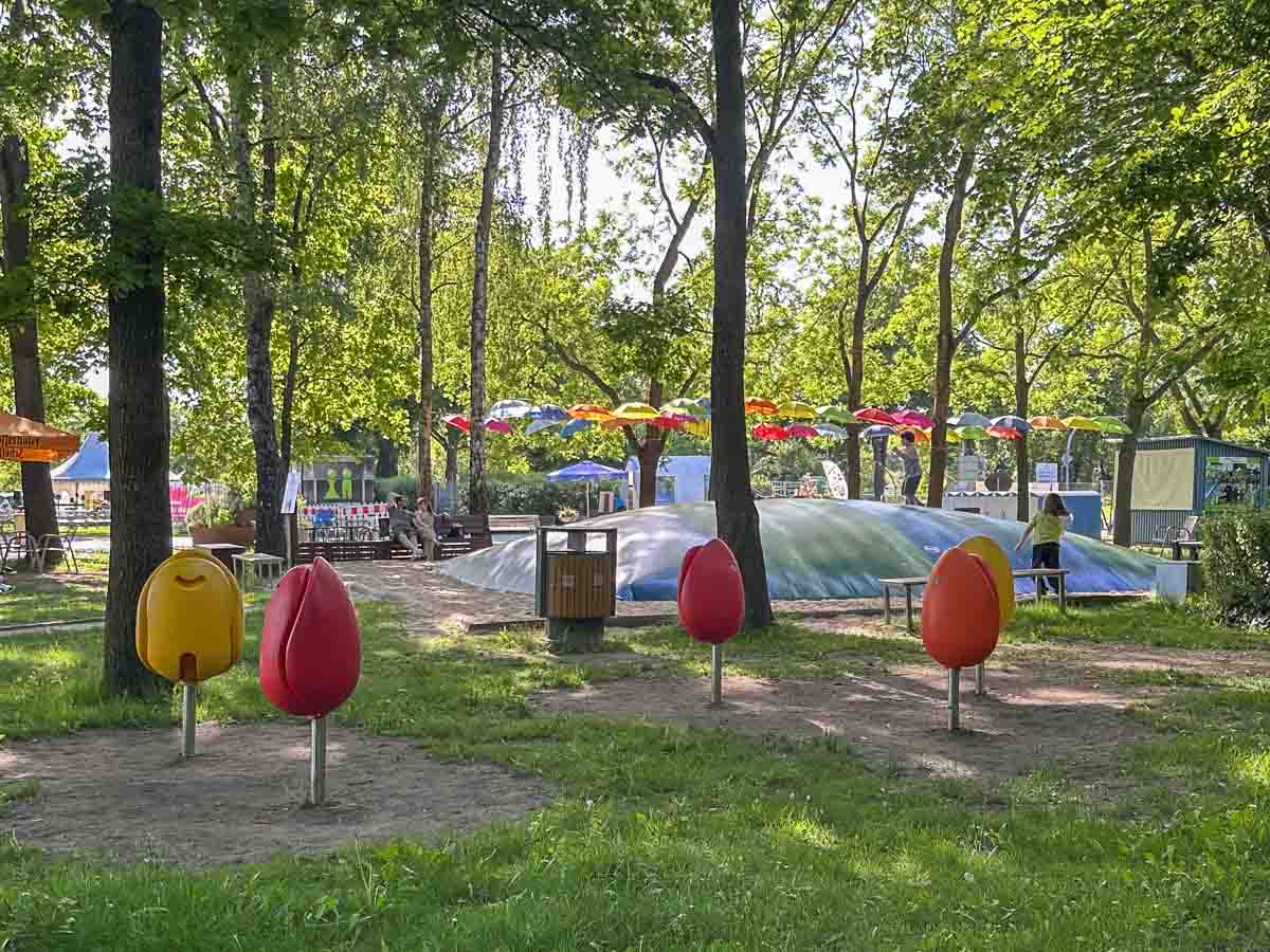 Auf einem Spielplatz in Havelland gibt es bunte Frühlingsschaukeln in Form von Früchten, eine große aufblasbare Kuppel, üppig grüne Bäume, Rasenflächen und Bänke. Über den Köpfen hängen bunte Sonnenschirme, während im Hintergrund Menschen sitzen und spielen.