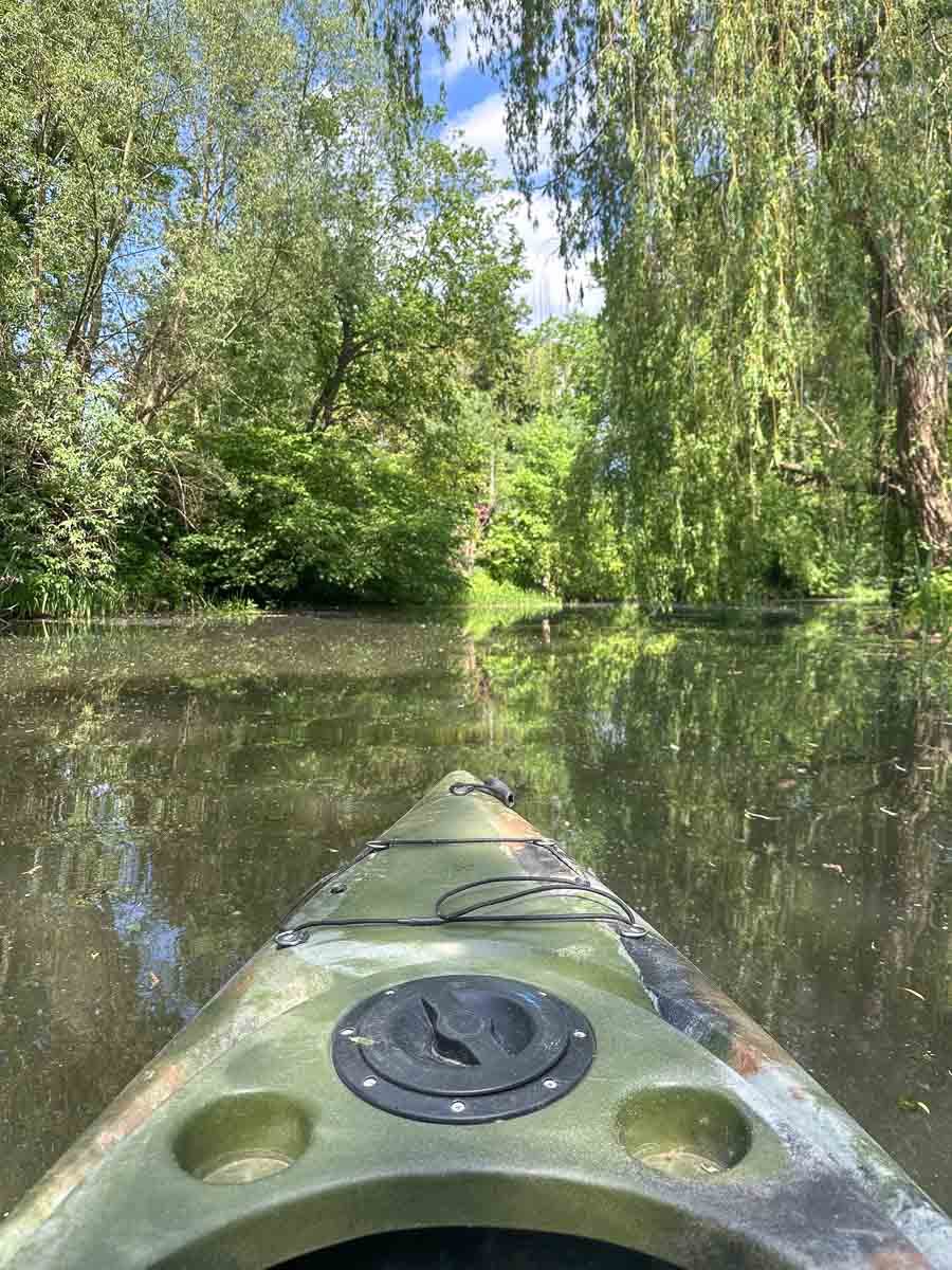 Blick aus einem Kajak auf einen ruhigen, schmalen Fluss im Havelland, umgeben von üppig grünen Bäumen und herabhängenden Ästen unter einem teilweise bewölkten Himmel. Im Vordergrund ist der Bug des Kajaks zu sehen.
