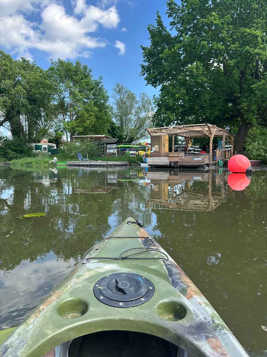 Ein grünes Kajak auf einem ruhigen Fluss im Havelland, das sich einem Uferbereich mit Holzkonstruktionen, Bäumen, Booten und großen roten Bojen unter einem teilweise bewölkten blauen Himmel nähert.