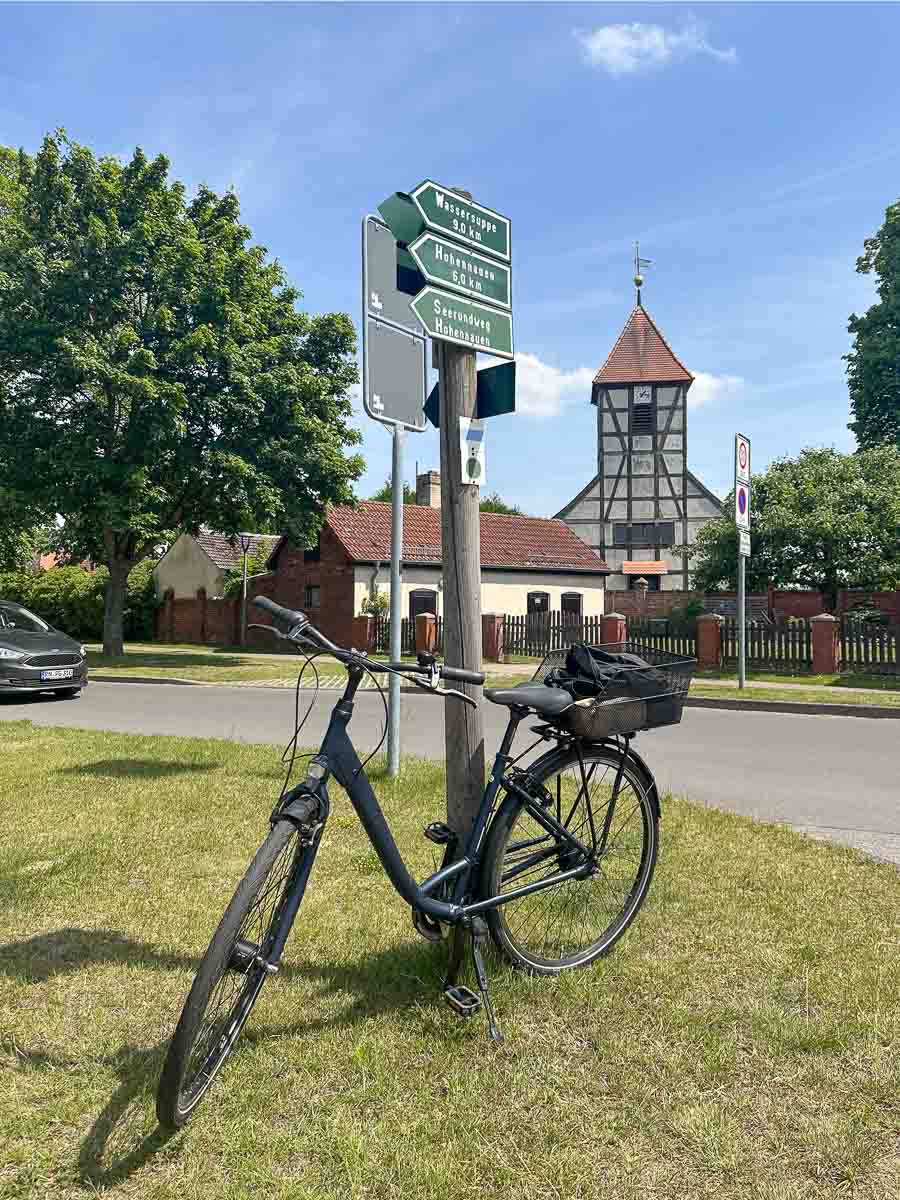 In Semlin im Westhavelland steht ein schwarzes Fahrrad an einem Wegweiser mit grünen Richtungsschildern. Im Hintergrund sind unter blauem Himmel Häuser, Bäume, ein geparktes Auto und eine Fachwerkkirche mit Uhrturm zu sehen.