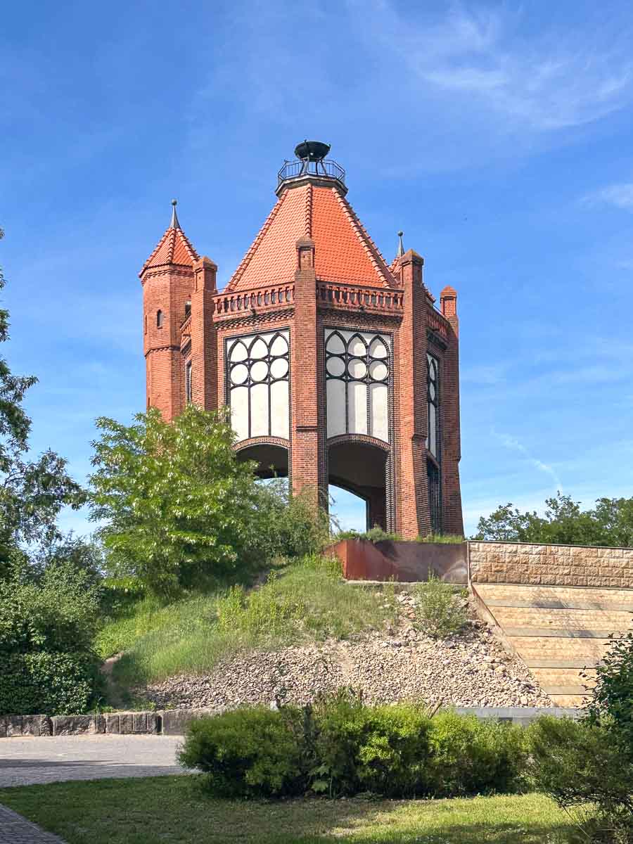 Der Bismarckturm, ein historischer Wasserturm aus rotem Backstein mit Bogenfenstern und einem steilen, gedeckten Dach steht auf einem grasbewachsenen Hügel in Rathenow im Havelland unter blauem Himmel, umgeben von üppigem Grün und einer Stützmauer aus Stein.
