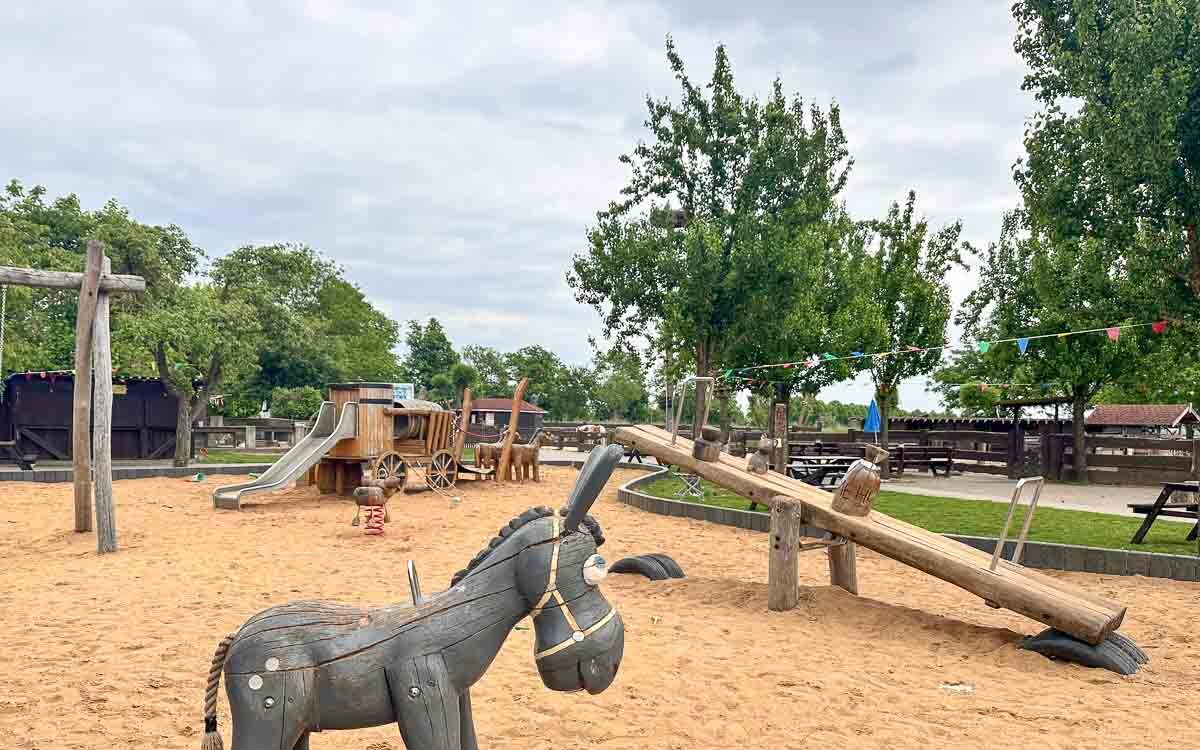 Auf einem Sandspielplatz bei Ribbeck im Havelland stehen eine Eselsskulptur aus Holz, eine Wippe, eine Rutsche und ein Piratenschiff. Im Hintergrund sind Bäume und Picknicktische unter einem bewölkten Himmel zu sehen.