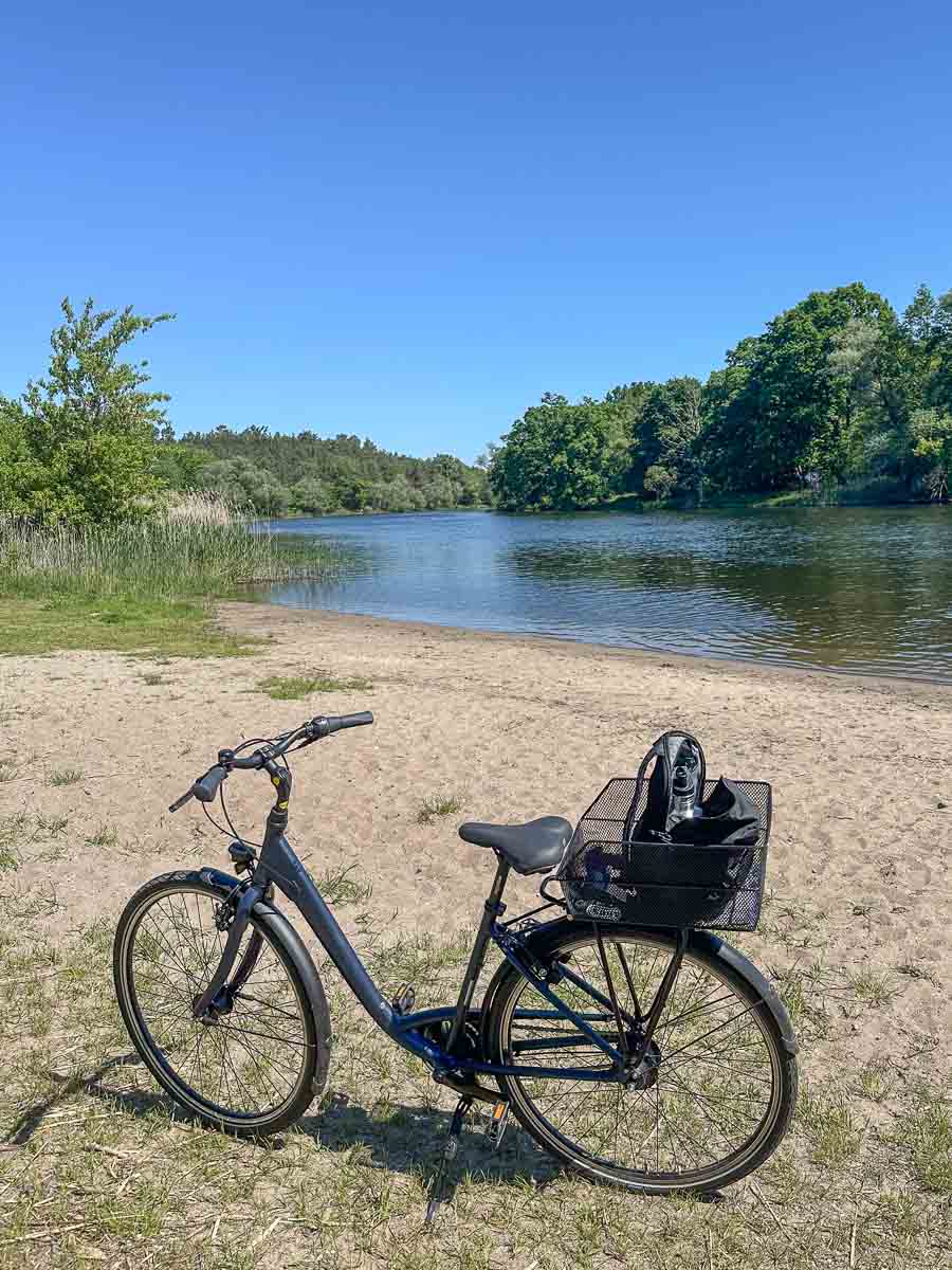 Ein schwarzes Fahrrad mit einem Korb steht auf sandigem Boden in der Nähe eines ruhigen Flusses im Westhavelland, umgeben von grünen Bäumen unter einem strahlend blauen Himmel.