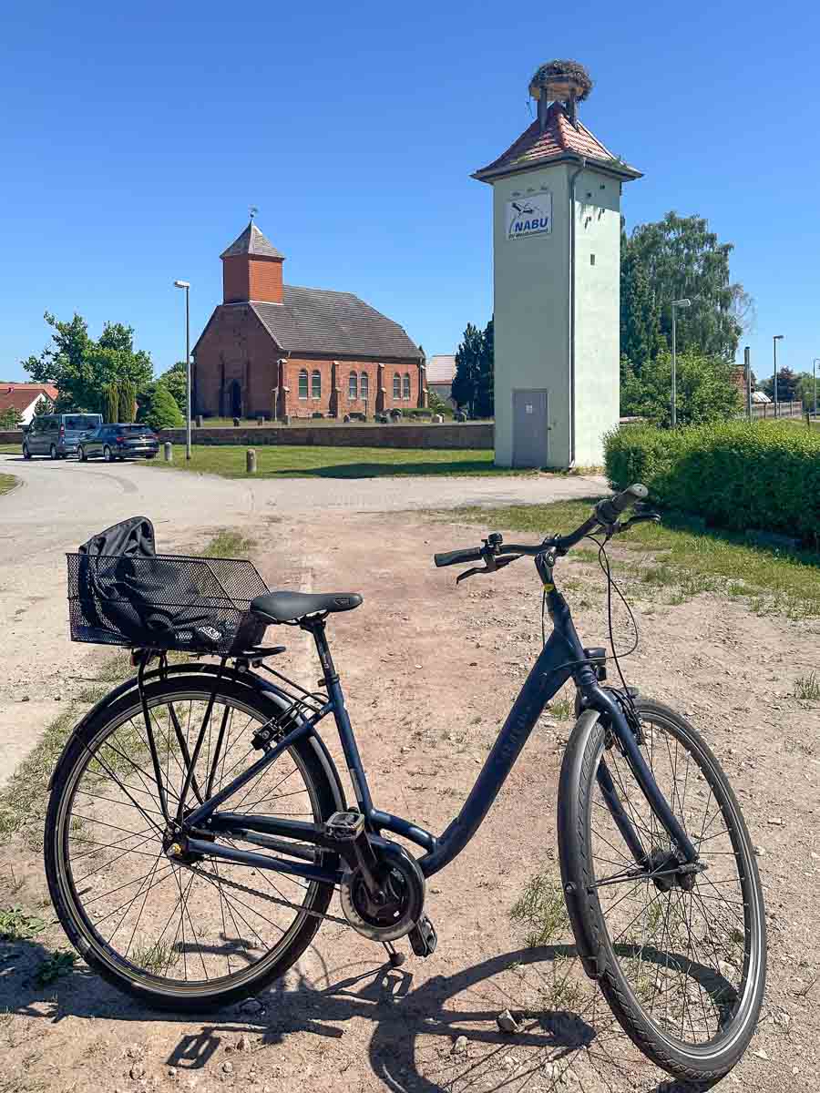 Ein schwarzes Fahrrad mit einem Korb hinten steht auf einem Feldweg in Havelland. Im Hintergrund ragen unter dem strahlend blauen Himmel ein grüner Turm mit einem Storchennest und eine Kirche aus rotem Backstein empor.