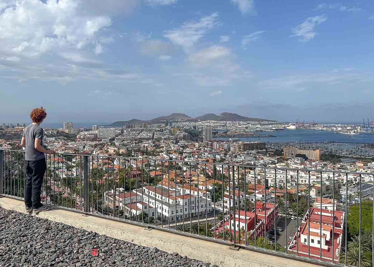 Ein Teenager mit lockigem Haar steht an einem Geländer und blickt auf die atemberaubende Stadtlandschaft von Las Palmas de Gran Canaria mit ihren Gebäuden mit roten Dächern, dem geschäftigen Hafen, dem Meer und den fernen Bergen unter einem teilweise bewölkten Himmel.