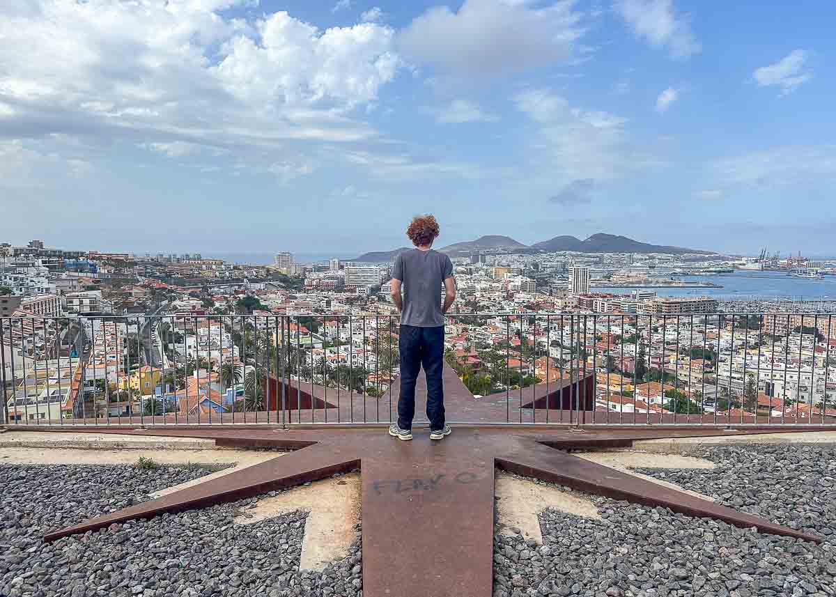 Ein Teenager mit lockigem Haar steht auf einem großen Metallstern und blickt unter einem teilweise bewölkten Himmel auf die Stadtlandschaft von Las Palmas de Gran Canaria mit ihren Gebäuden, der Bucht, den Schiffen und den Bergen – der Mirador de Agustin Castillo ist ein idealer Ort für alle, die Geheimtipps suchen.