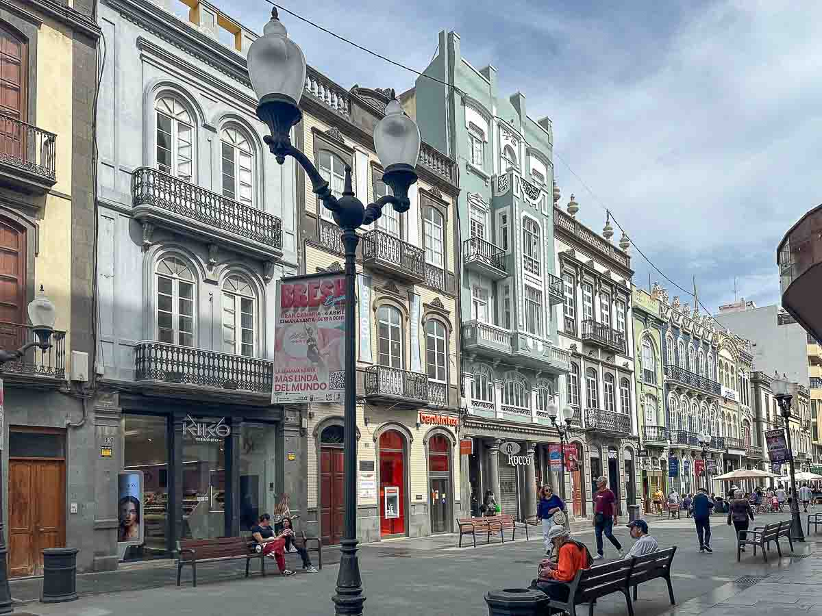 Die Calle Triana ist eine Fußgängerzone, in der Menschen auf Bänken sitzen und spazieren gehen. Historische Gebäude mit verzierten Fassaden und Balkonen säumen die Straße und bilden eine der reizvollen Sehenswürdigkeiten in Las Palmas de Gran Canaria. Unter einem teilweise bewölkten Himmel sind Straßenlaternen und Geschäfte zu sehen