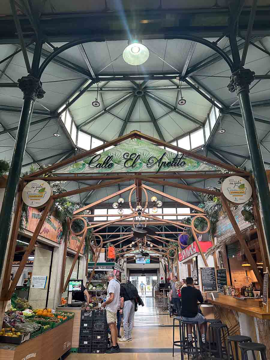 Die Markthalle Mercado del Puerto mit gewölbten Dachstützen, Obst- und Gemüseständen auf der linken Seite, einer Bar auf der rechten Seite und einem Schild mit der Aufschrift „Calle El Apetito“ über dem Durchgang. Mehrere Menschen sind beim Einkaufen oder sitzen an der Bar – eine von vielen authentischen Sehenswürdigkeiten in Las Palmas de Gran Canaria für Familien.