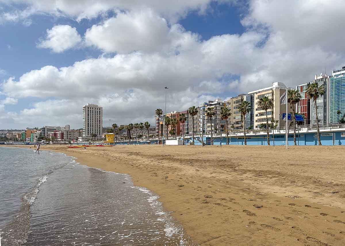 Der breite Sandstrand der Playa Alcaravaneras mit sanften Wellen, ein paar Menschen, die am Ufer entlangspazieren, und eine Reihe moderner Gebäude und Palmen, die unter einem teilweise bewölkten Himmel die Strandpromenade säumen –  ein idealer Ort in Las Palmas de Gran Canaria mit Kindern