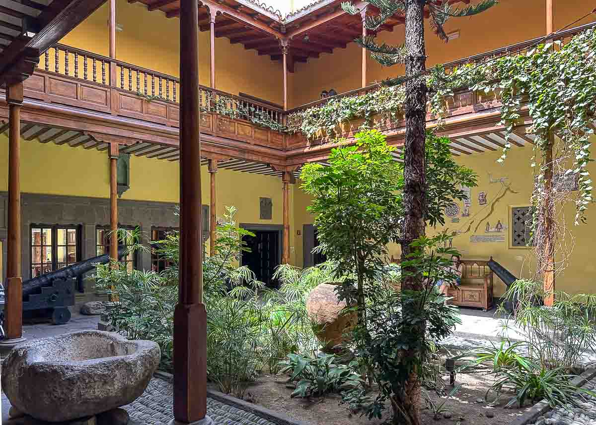 Ein sonniger Innenhof mit gelben Wänden, einem Holzbalkon, üppigen Grünpflanzen, einem Steinbecken, Kanonen und einem kunstvollen Wandgemälde; im Stil traditioneller spanischer oder kolonialer Architektur: Die Casa de Colon ist  eine der reizvollsten Sehenswürdigkeiten in Las Palmas de Gran Canaria.