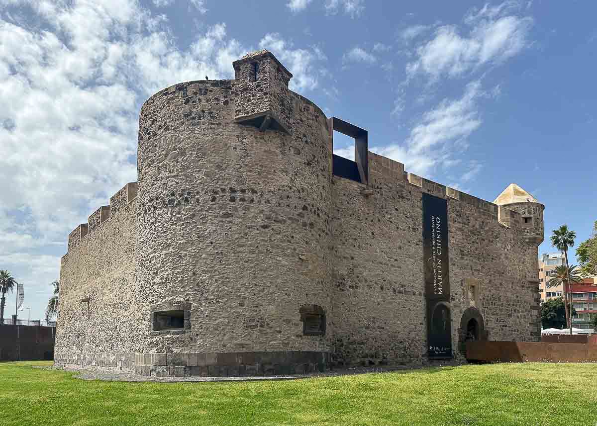 Unter einem teilweise bewölkten Himmel erhebt sich auf einer grasbewachsenen Wiese eine große historische Steinfestung mit runden Türmen; am Eingang hängt ein Banner, und im Hintergrund sind Palmen zu sehen – einer der Geheimtipps in Las Palmas de Gran Canaria.