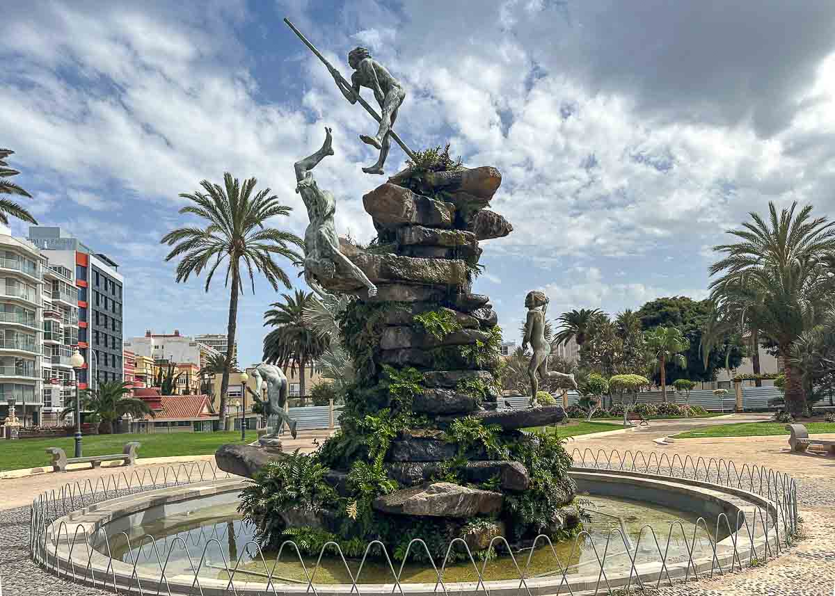 Ein Springbrunnen mit Bronzestatuen von Menschen, die auf eine felsige zentrale Struktur klettern und von ihr herunterspringen, umgeben von Wasser, Palmen und Stadtgebäuden im Hintergrund – eine der Sehenswürdigkeiten in Las Palmas de Gran Canaria, die man unbedingt gesehen haben muss. Lassen Sie sich diesen Ort nicht entgehen, wenn Sie auf der Suche nach unvergesslichen Tipps sind, um die künstlerische Seite der Stadt zu erkunden.