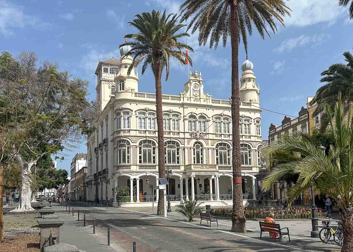 Historisches weißes Gebäude mit verzierten Türmen, Palmen und einer Fußgängerzone davor. Das Gabinete Literario, dieses beeindruckende Wahrzeichen im Herzen von Las Palmas de Gran Canaria zählt zu den Top-Sehenswürdigkeiten der Stadt – ein lohnender Besuch für alle, die auf der Suche nach Insidertipps sind. Über dem Gebäude ist der Himmel klar, am Eingang sind einige Menschen und ein Radfahrer zu sehen.