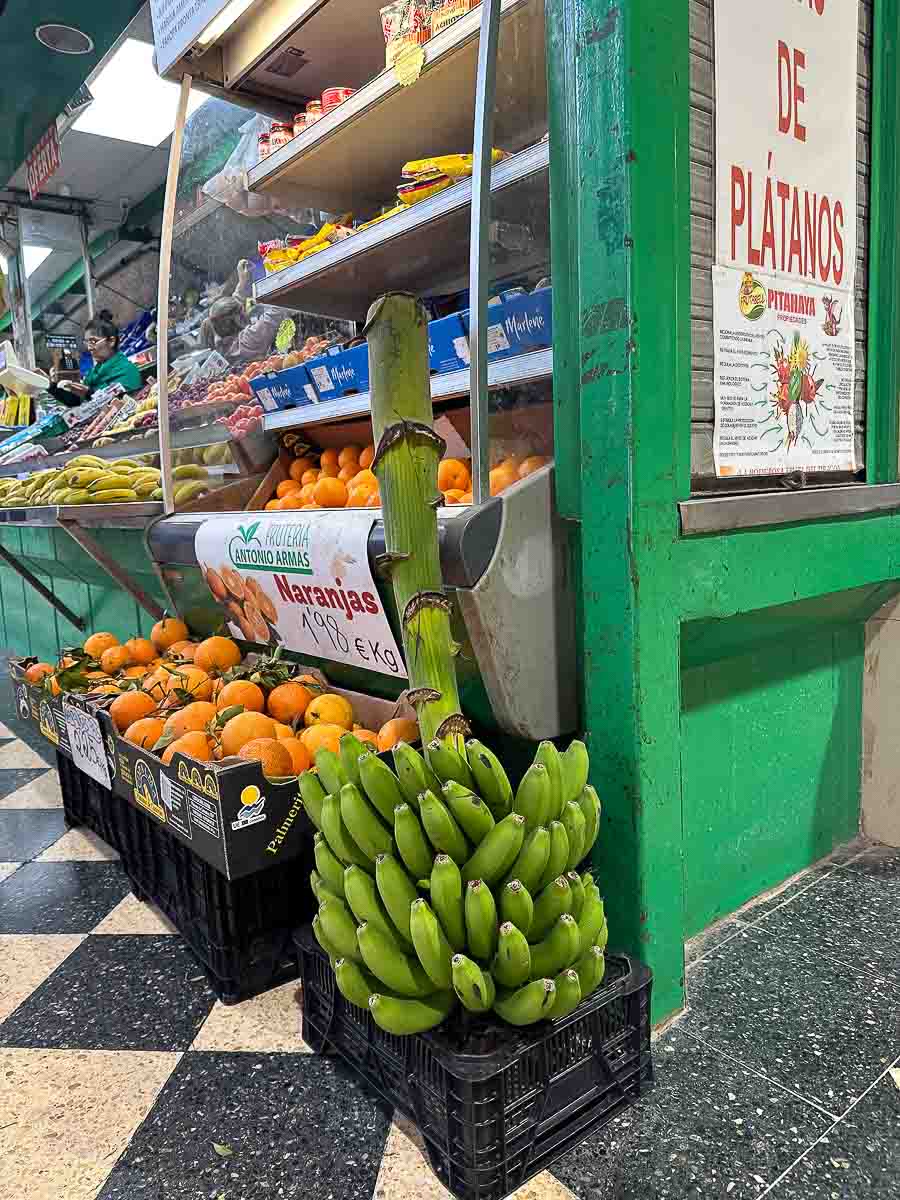 In der Markthalle Mercado Central in Las Palmas de Gran Canaria steht ein Bund grüner Kochbananen aufrecht auf einer Kiste, umgeben von Orangen, Obst und einem Schild mit der Aufschrift „PLÁTANOS“. Der Boden ist mit schwarz-weiß karierten Fliesen ausgelegt – eine authentische Kulisse für alle, die nach Tipps suchen oder die lokalen Sehenswürdigkeiten erkunden möchten.