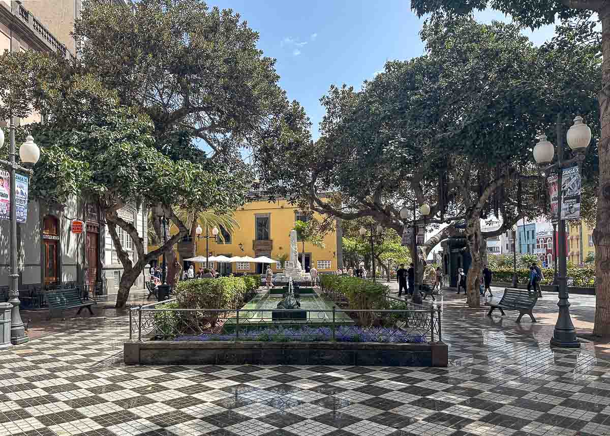 Ein Stadtplatz mit gekacheltem schwarz-weißen Boden, Bänken, Bäumen, einem großen Springbrunnen in der Mitte und Spaziergängern; im Hintergrund sind unter einem teilweise bewölkten Himmel ein gelbes Gebäude und Sitzgelegenheiten eines Straßencafés zu sehen – die Plaza de las Ranas ist ein perfekter Ort für Familien in Las Palmas de Gran Canaria.