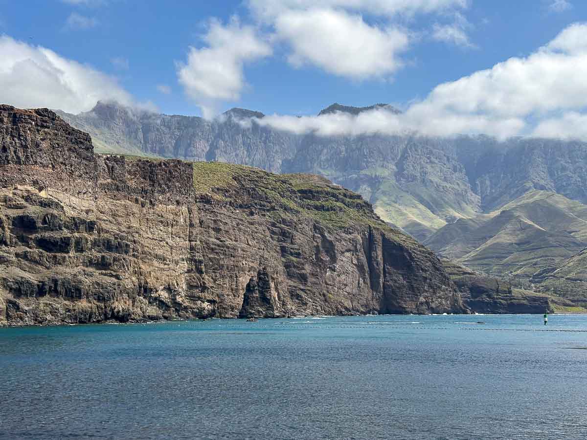 Felsige Klippen ragen aus dem blauen Wasser empor, im Hintergrund sind üppig grüne Flecken und schroffe Berge unter einem teilweise bewölkten Himmel zu sehen – die Kulisse der Westküste bei Puerto de las Nieves, einem der beliebten Gran Canaria Ausflugsziele.