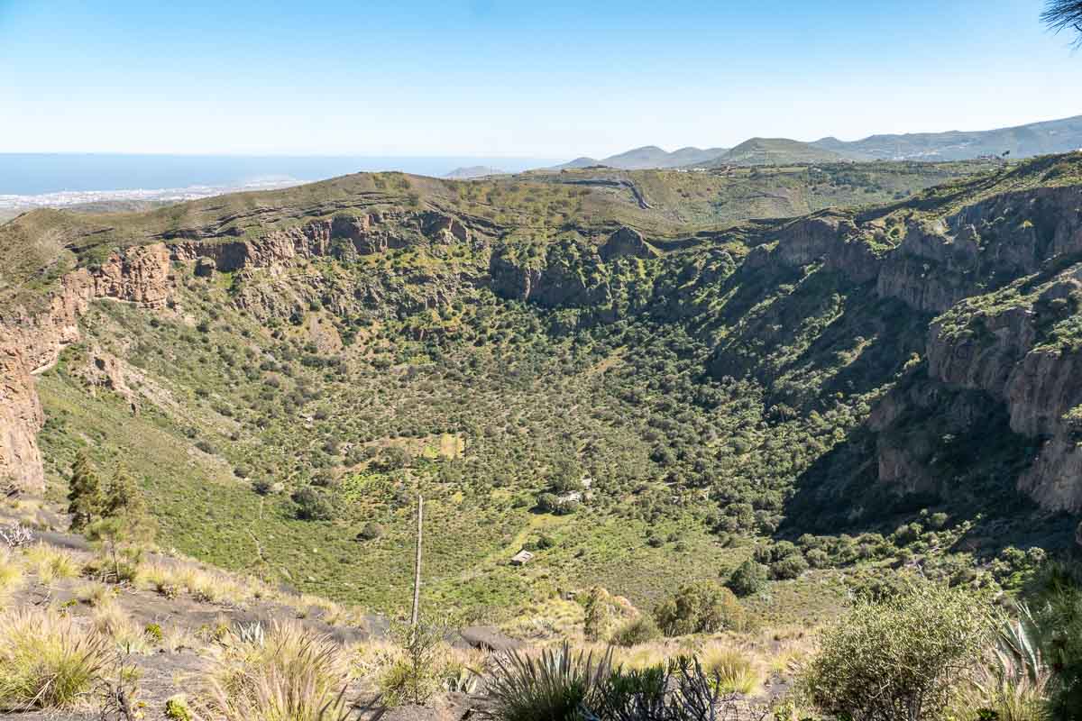 Die Caldera de Bandama ist ein weitläufiger, von grüner Vegetation bedeckter Vulkankrater, umgeben von felsigen Klippen und mit fernen Hügeln im Hintergrund unter einem strahlend blauen Himmel und ein beliebtes Ziel für Ausflüge von Las Palmas de Gran Canaria