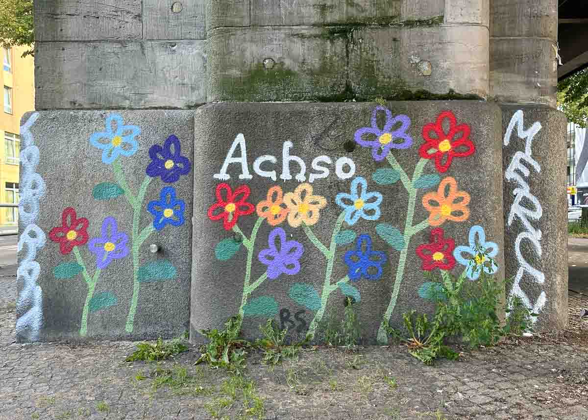 Auf einer Betonmauer im Freien in der Nähe des Urban Nation Museums sind bunte Blumen sowie die Wörter „Achso“ und „Merci“ aufgesprüht; am Fuß der Mauer wächst etwas Gras.