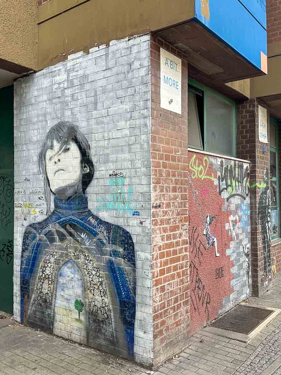 An einer Eckwand in der Nähe des Urban Nation Berlin ist ein Wandbild zu sehen, das eine Person mit dunklem Haar und geschlossenen Augen zeigt; es ist von Graffiti und Tags umgeben. Der untere Teil des Wandbilds ist mit farbenfrohen, abstrakten Mustern verziert.