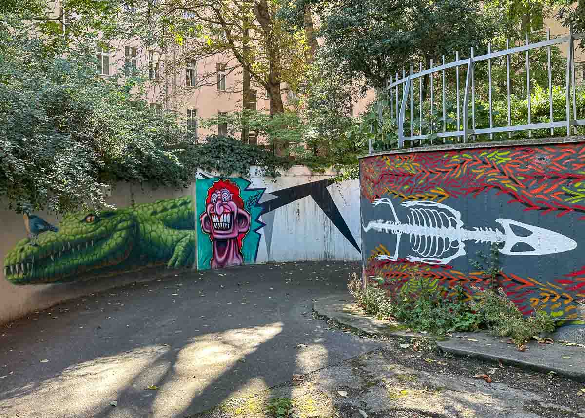 Farbenfrohe Streetart an zwei geschwungenen Wänden in der Nähe des Streetart Museum Berlin zeigt ein grünes Krokodil, eine karikaturhafte Figur mit rosa Gesicht und ein weißes Fischskelett vor einem leuchtend roten und gelben Hintergrund, umgeben von Bäumen und einem Metallzaun darüber.