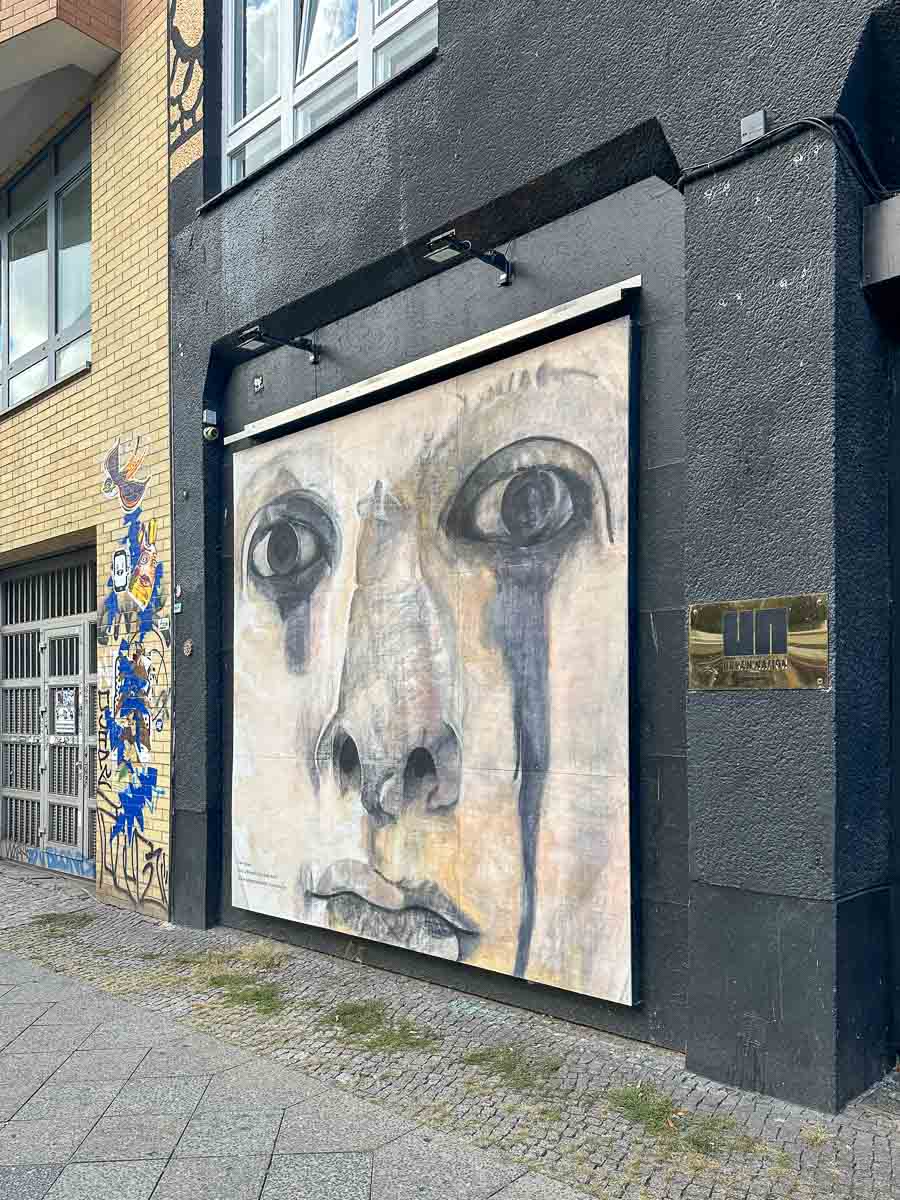 Großes Wandgemälde eines Gesichts mit tränenfeuchten Augen an der schwarzen Gebäudewand des Projektraums des Urban Nation Museum; daneben ist auf gelben Ziegeln ein farbenfrohes Wandgemälde mit abstrakten Motiven zu sehen.
