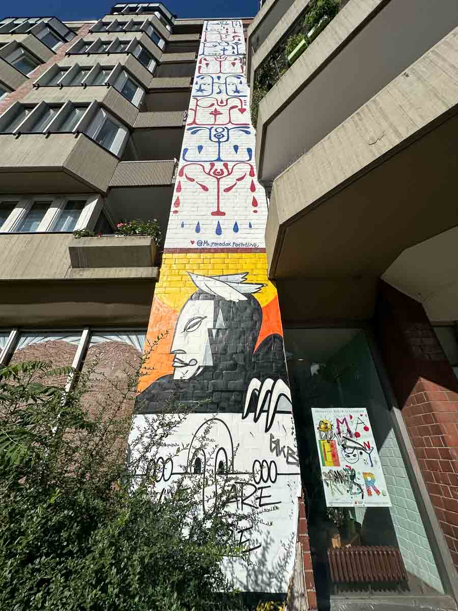 Ein großes Wandbild an einem Gebäude in der Nähe des Urban Nation Museums zeigt abstrakte Figuren, ein gekröntes Wesen und Graffiti am unteren Rand. Das Wandbild wird von Betonbalkonen und Grünflächen eingerahmt.