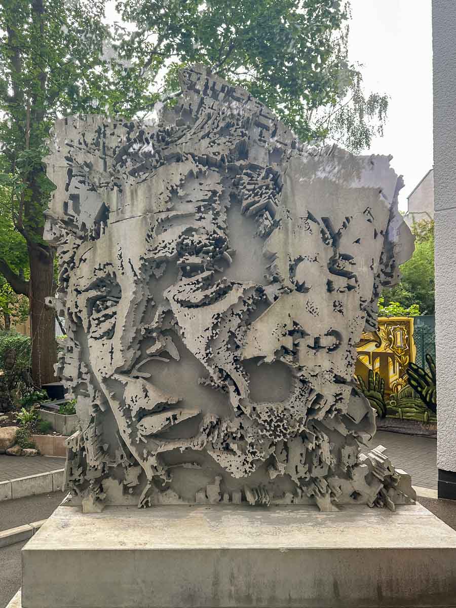 Übergroße Betonskulptur, die ein fragmentiertes menschliches Gesicht mit gezackten, unebenen Kanten darstellt; ausgestellt inmitten von Bäumen und Grünflächen im Außenbereich des Urban Nation Museum.